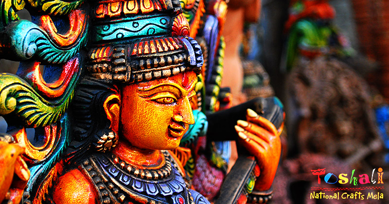 Una statua di legno colarata di profilo e in basso a destra la scritta Toshali National Craft Mela