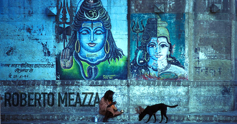 un sadhu con un cane davanti a un muro dipinto con shiva