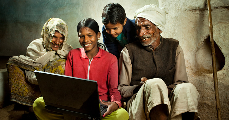Una ragazzina indiana mostra sorridente un portatile al nonno, fratello e mamma vestiti con i vestiti tipici degli indiani delle campagne del nord